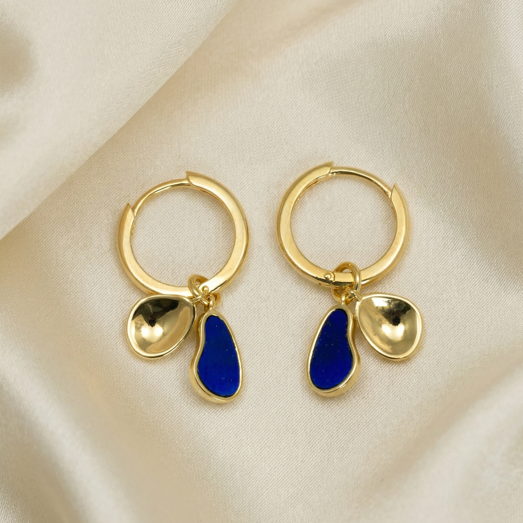 Pair of gold vermeil hoop earrings each featuring two sculptural charms, one polished gold-tone and one with vibrant blue lapis inlay, displayed on soft neutral fabric.