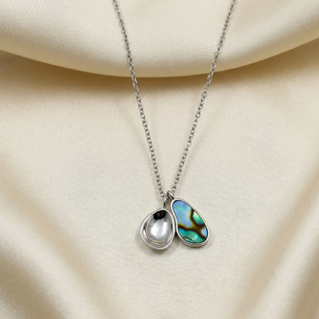 Sterling silver necklace featuring a double pendant with a polished silver teardrop and an iridescent abalone charm, displayed on a smooth cream fabric background.
