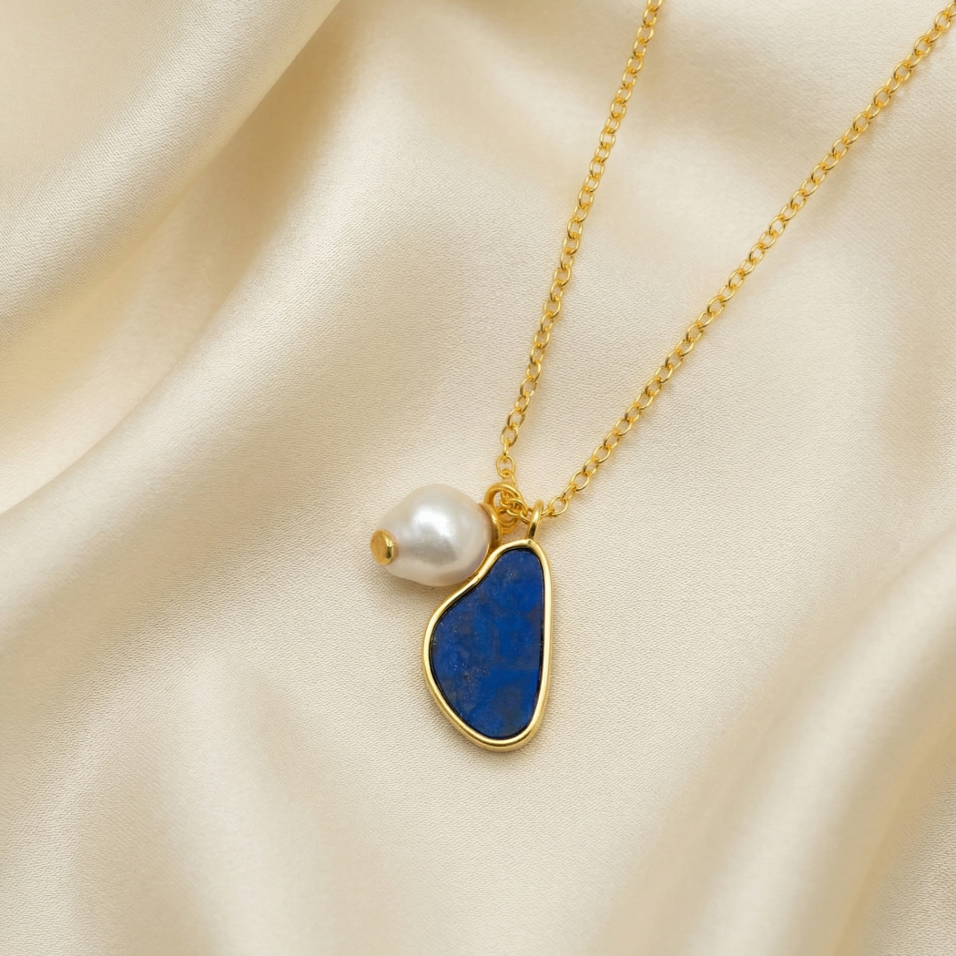 Gold vermeil necklace featuring a sculptural blue lapis pendant and an irregular baroque pearl, displayed on soft cream satin to highlight its elegant design.