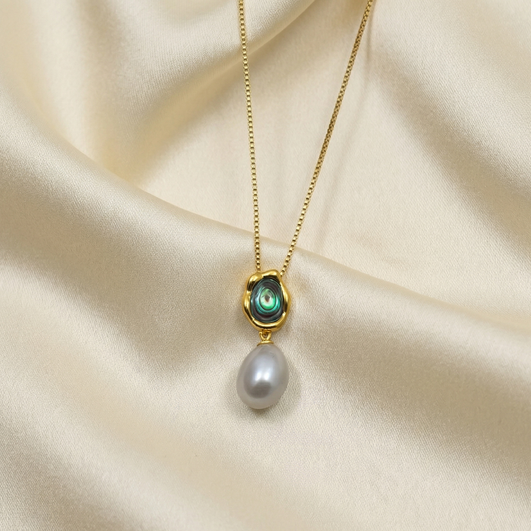 Gold vermeil necklace featuring a sculptural abalone shell detail above a lustrous white pearl drop pendant, displayed on a smooth cream fabric background.