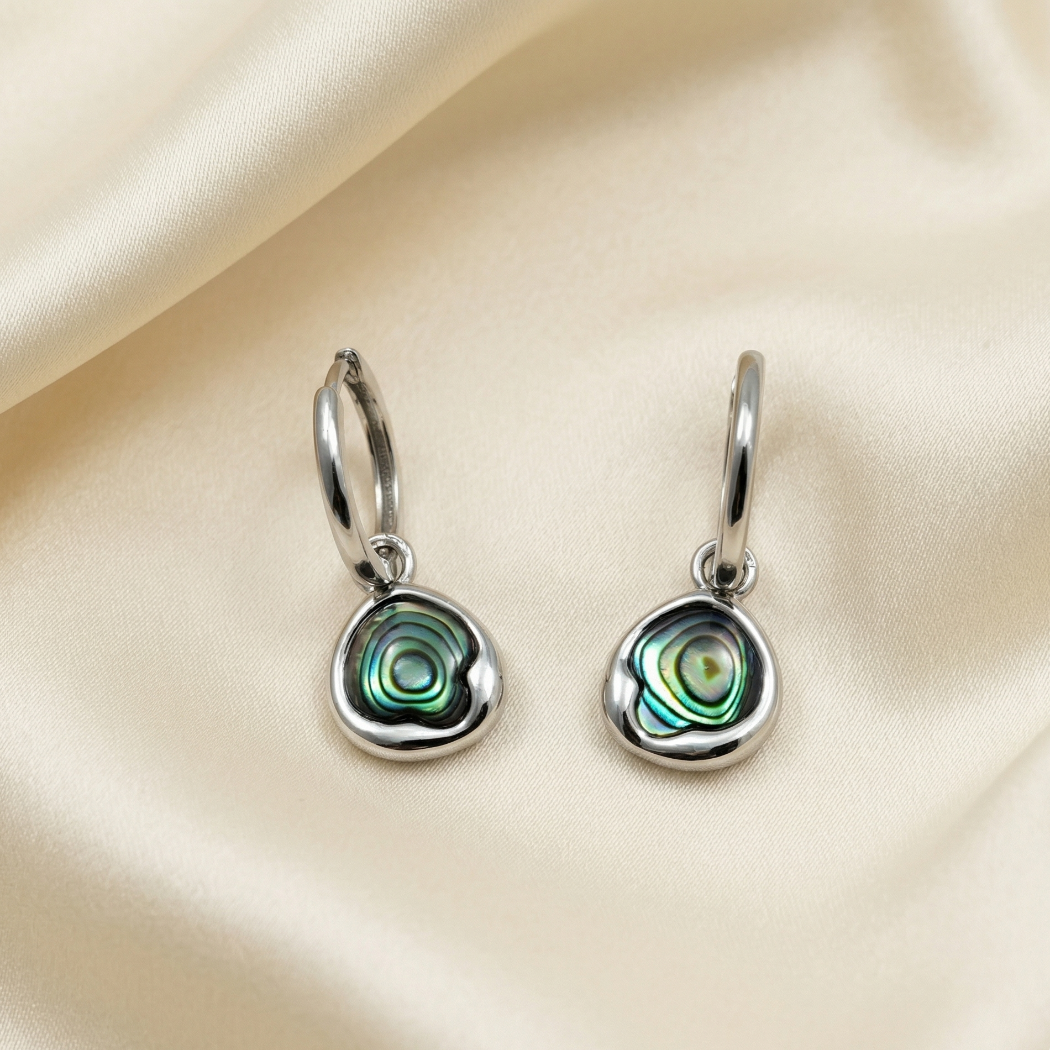 Pair of polished sterling silver hoop earrings featuring uniquely shaped abalone shell charms with iridescent blue-green and black swirls, displayed on smooth cream fabric.