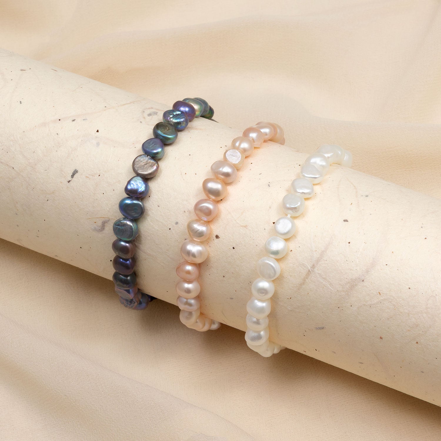 Three stretch bracelets featuring lustrous white, peach, and dark iridescent freshwater pearls, arranged on a neutral fabric background.