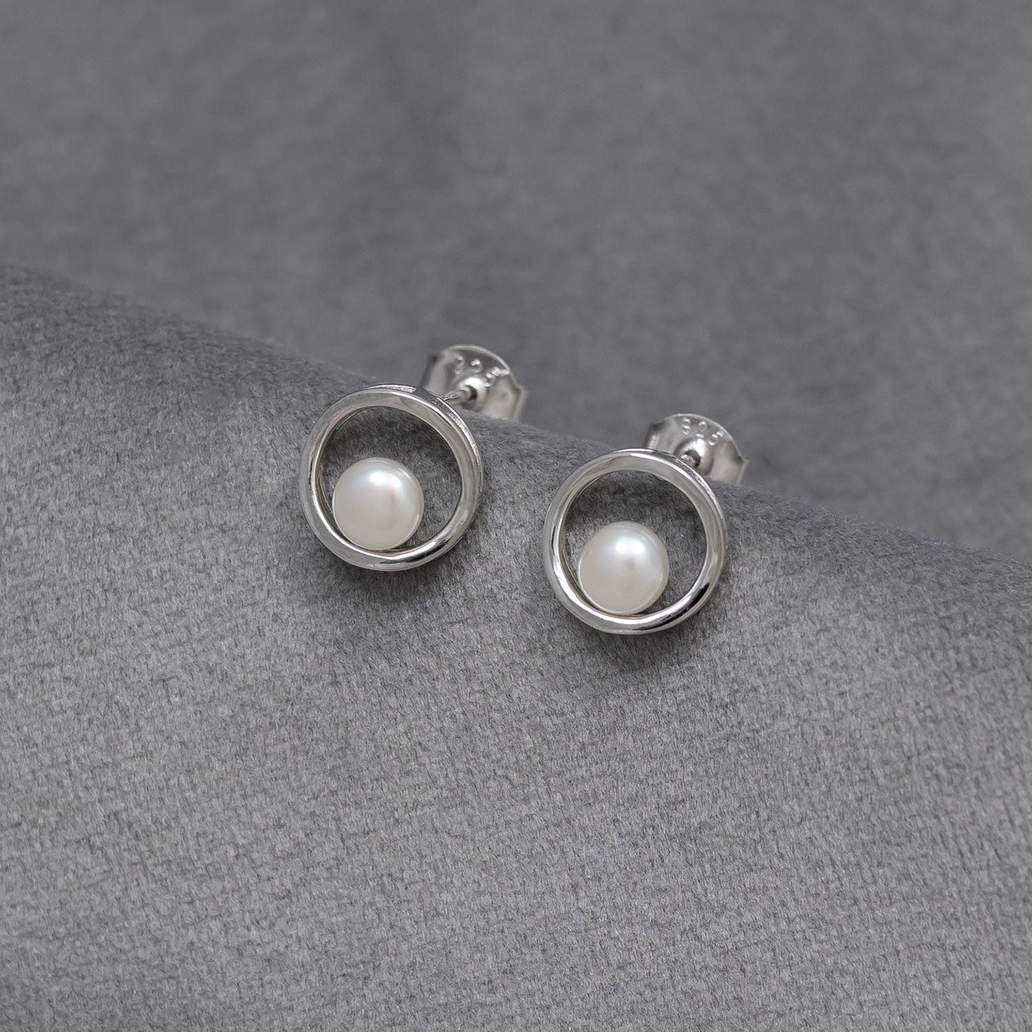 Pair of sterling silver stud earrings featuring a simple open circle design encasing a single luminous pearl, displayed on a soft grey fabric background.