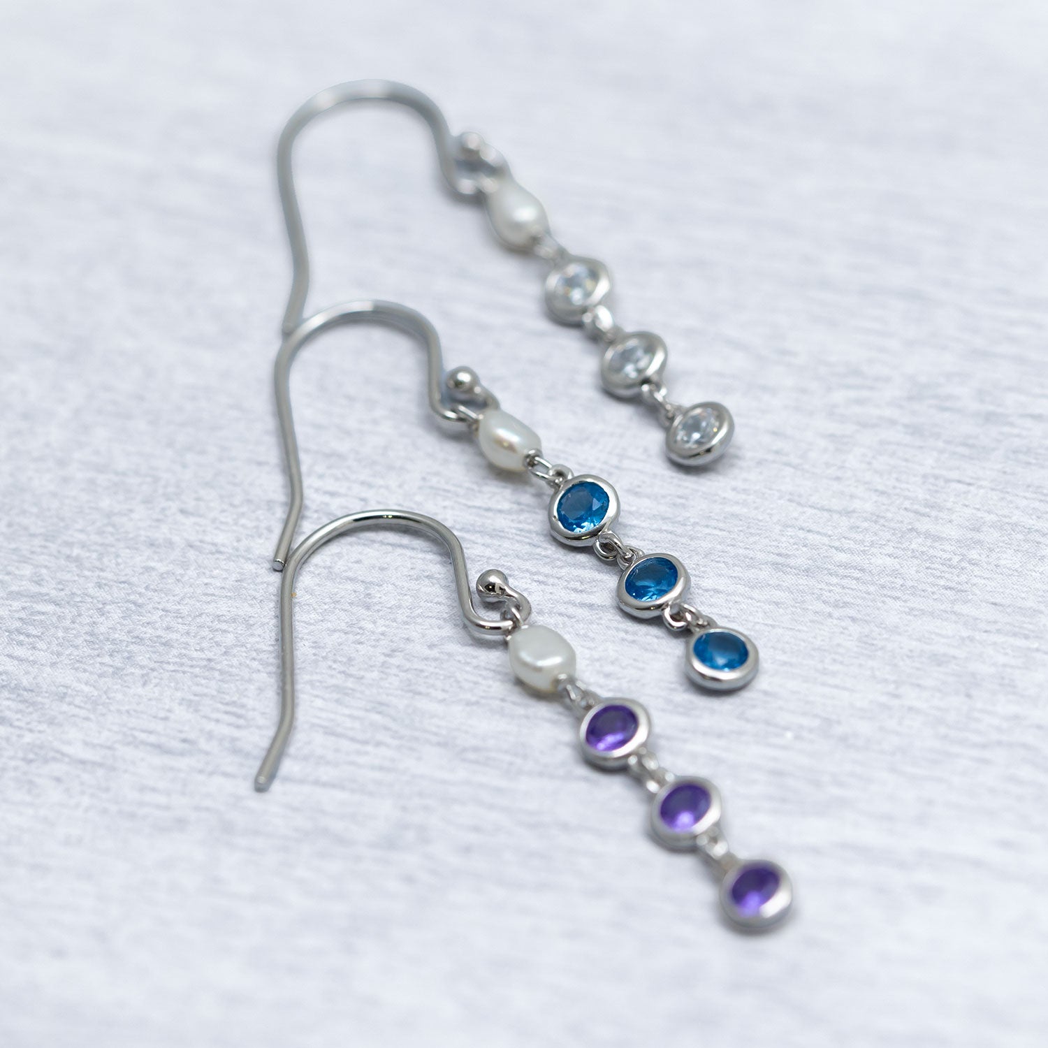 Three pairs of sterling silver drop earrings featuring delicate freshwater pearls paired with blue, purple, and clear cubic zirconia stones, highlighting a sculptural contemporary design.