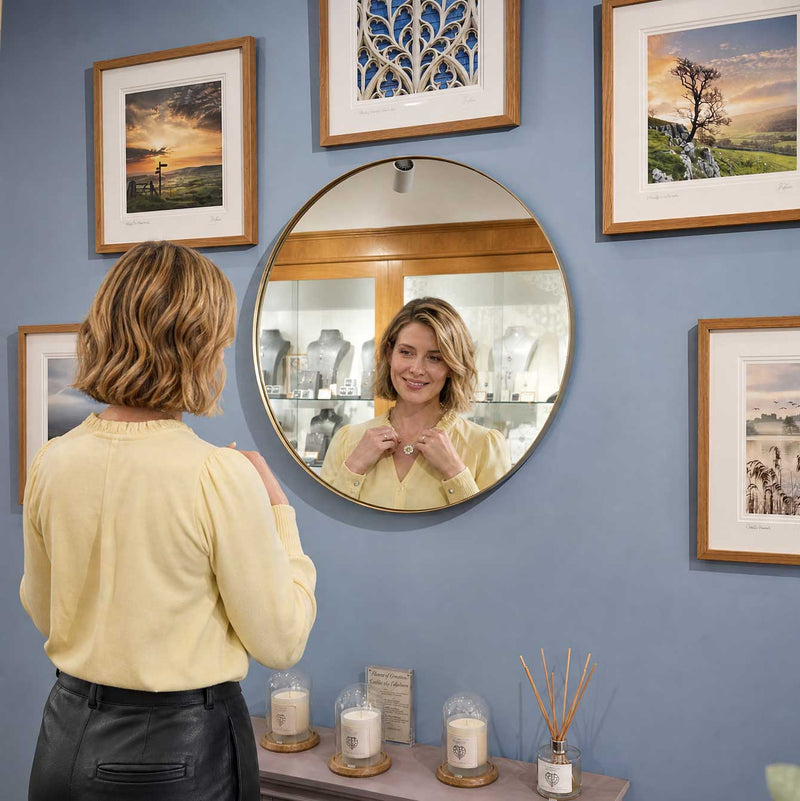 A woman with shoulder-length blonde hair wearing a pale yellow blouse and black trousers adjusts a necklace while looking into a round wall mirror above a shelf with candles and reed diffusers, surrounded by framed landscape photographs on a blue wall.