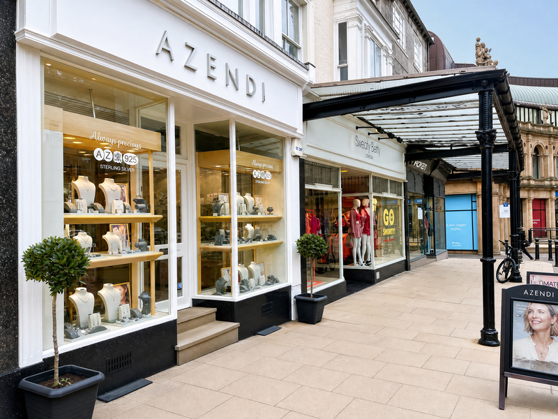 A street view of a contemporary jewellery boutique with large glass windows displaying sterling silver pieces on white busts, flanked by small potted trees under a glass canopy.