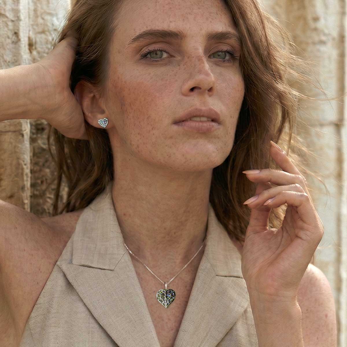 Portrait of a woman with natural makeup wearing a sleeveless beige top, showcasing a sterling silver heart-shaped pendant necklace and matching stud earrings with intricate detailing.