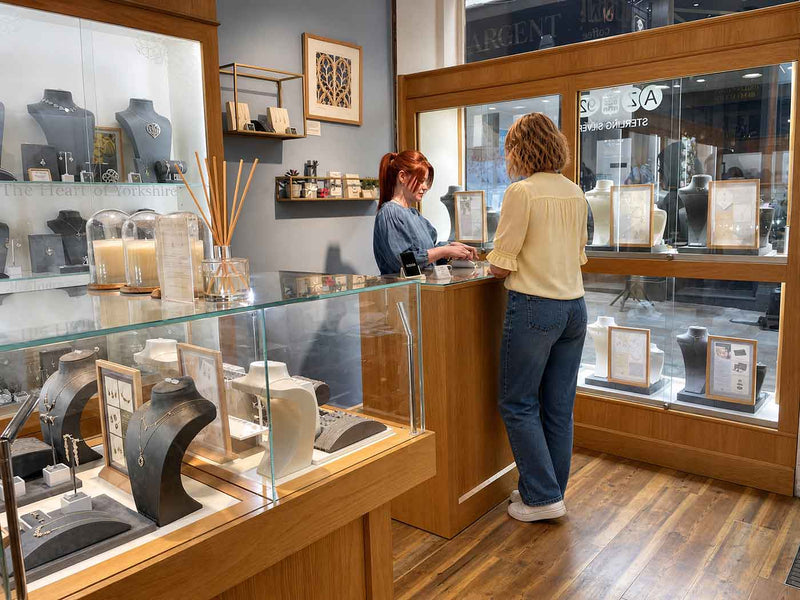 Inside a stylish boutique with wooden floors and display cases featuring sterling silver jewellery, a customer in a pale yellow blouse is assisted by a staff member behind the counter.