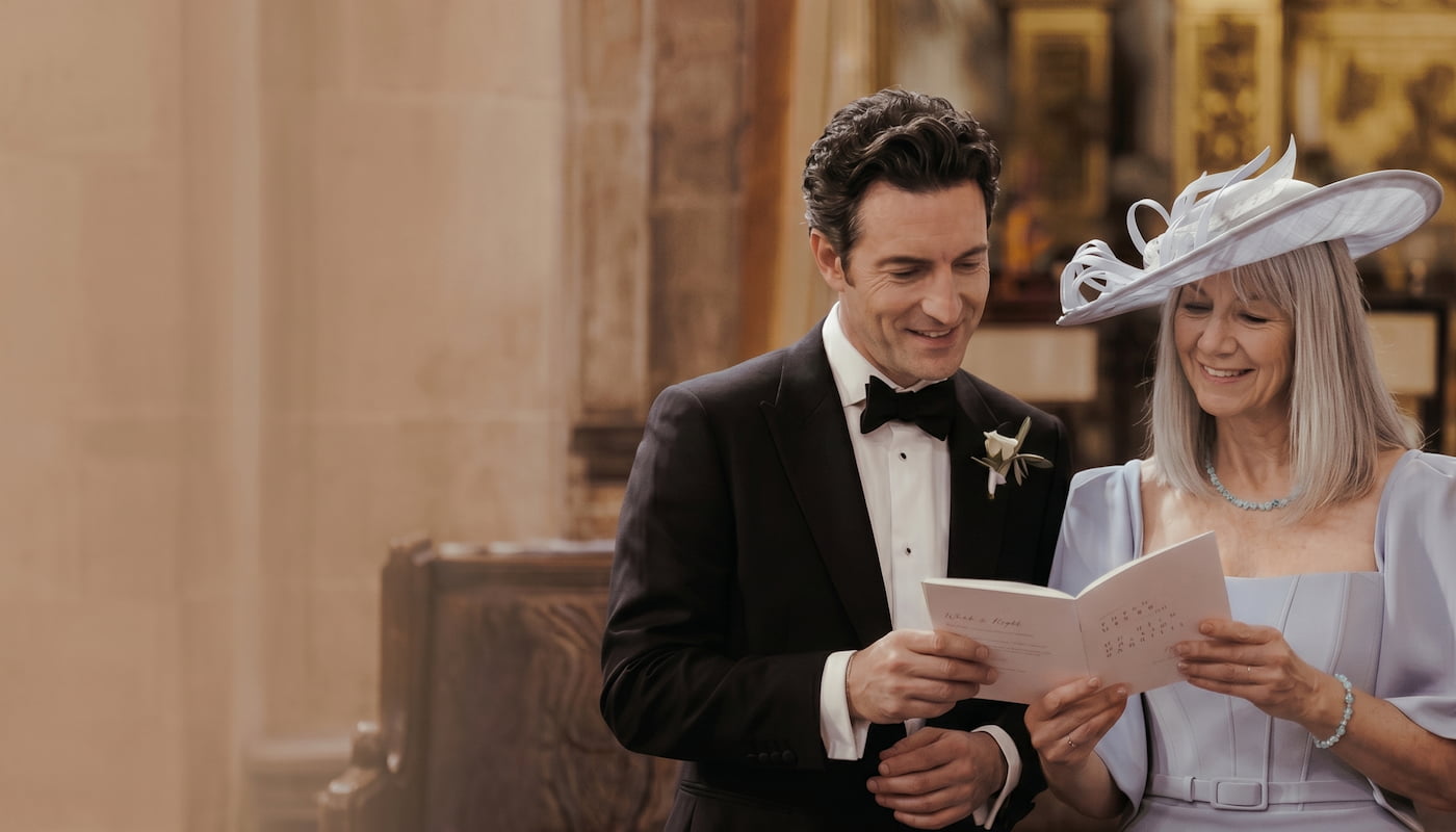 Groom in tuxedo and woman in formal attire reading an order of service together in a church.