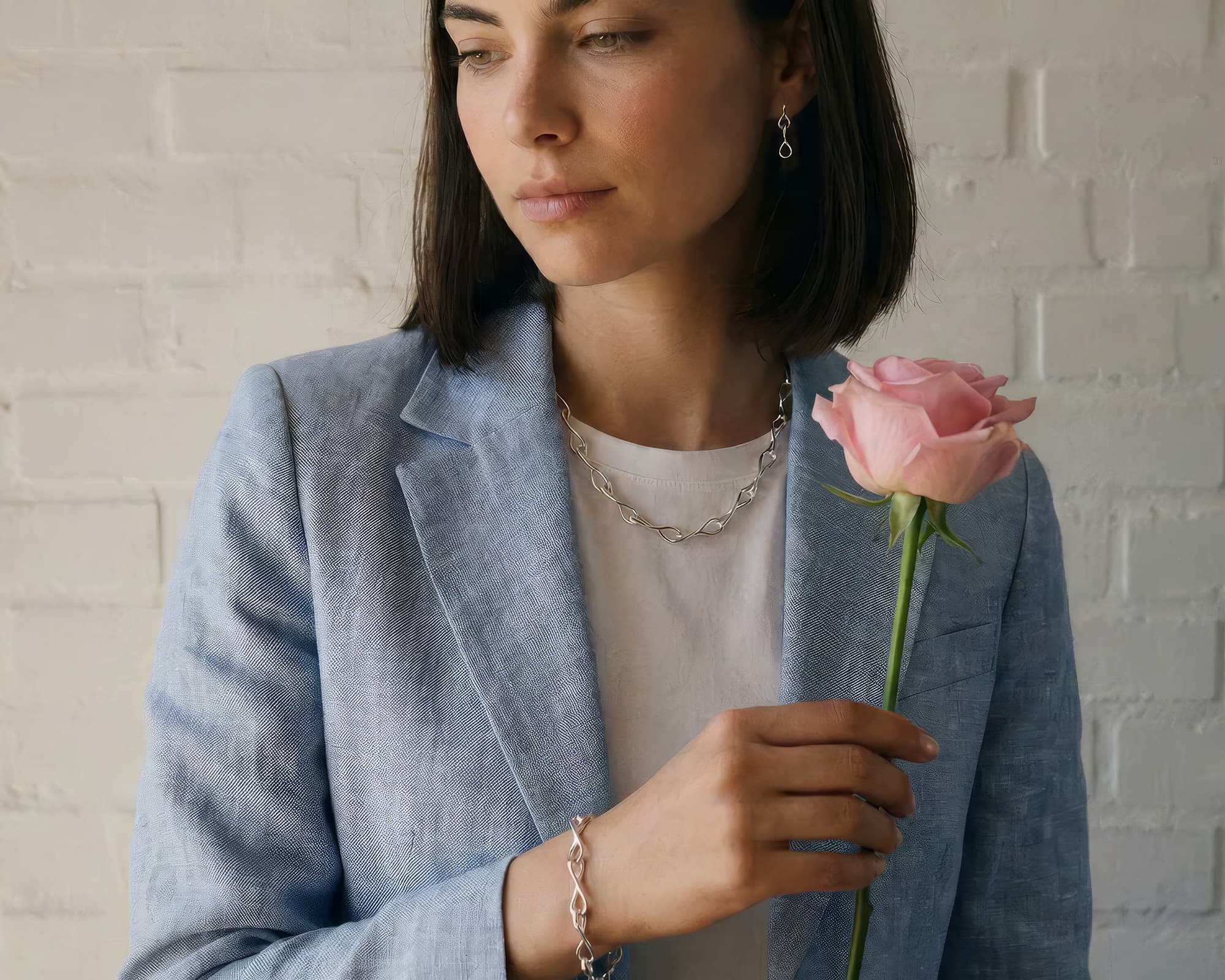 Woman holding a pink rose against a white brick wall wearing solid silver necklace from azendi