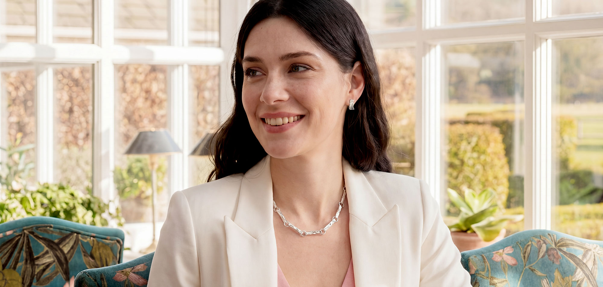 Woman in a white blazer sitting in a sunlit Yorkshire lounge with large windows. Wearing Malham Cove jewellery set.