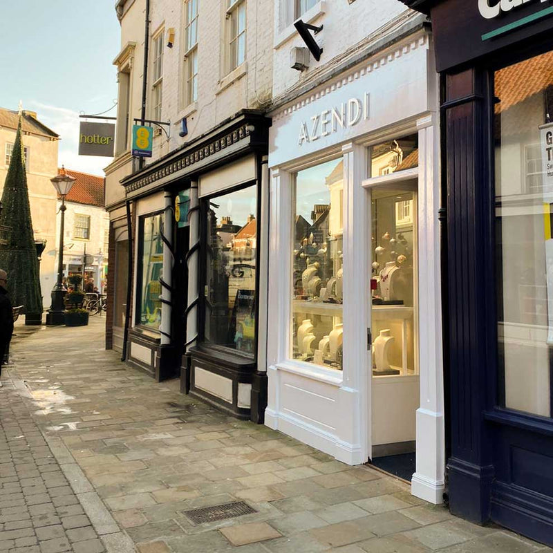 Street view of Toll Gavel with a white framed Azendi store, showcasing sterling silver jewellery in illuminated window displays during daylight.