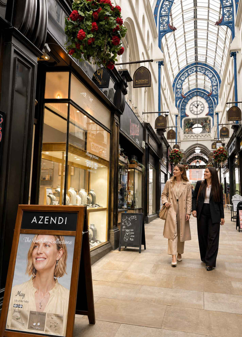 A bright, airy indoor shopping arcade with a high arched glass ceiling and decorative ironwork, featuring Azendi jewellery boutiques and two women walking along the polished walkway.