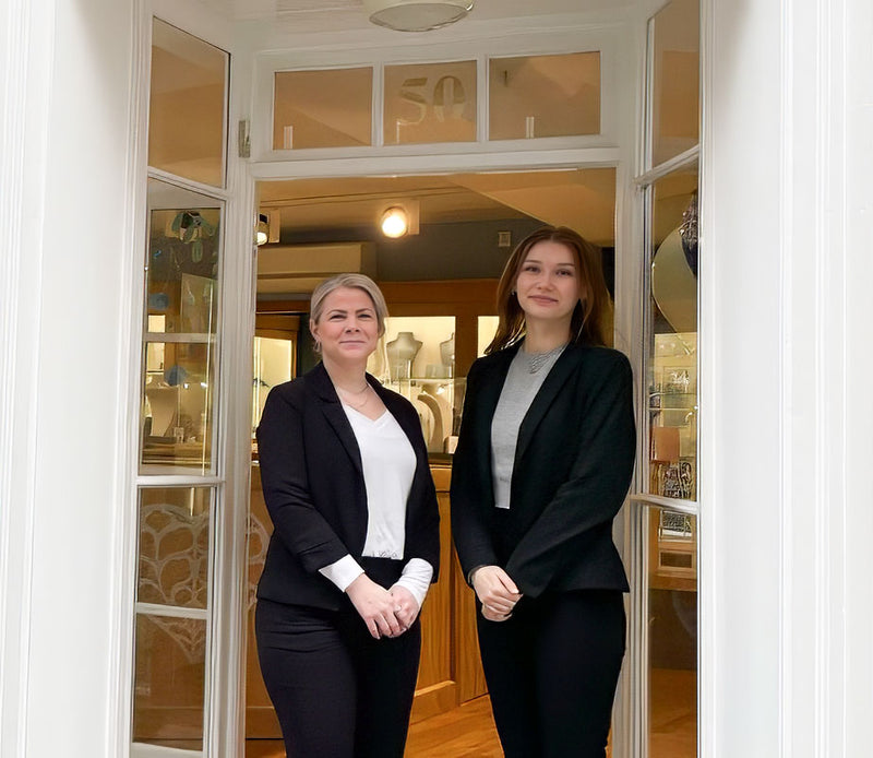 Two women dressed in professional black and white attire standing confidently in a bright doorway with display cases visible inside the elegant interior behind them.