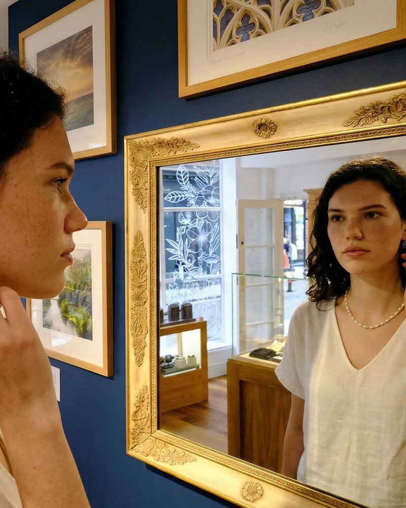 Woman looking at herself in a gold-framed mirror against a blue wall with framed pictures inside the Azendi York store