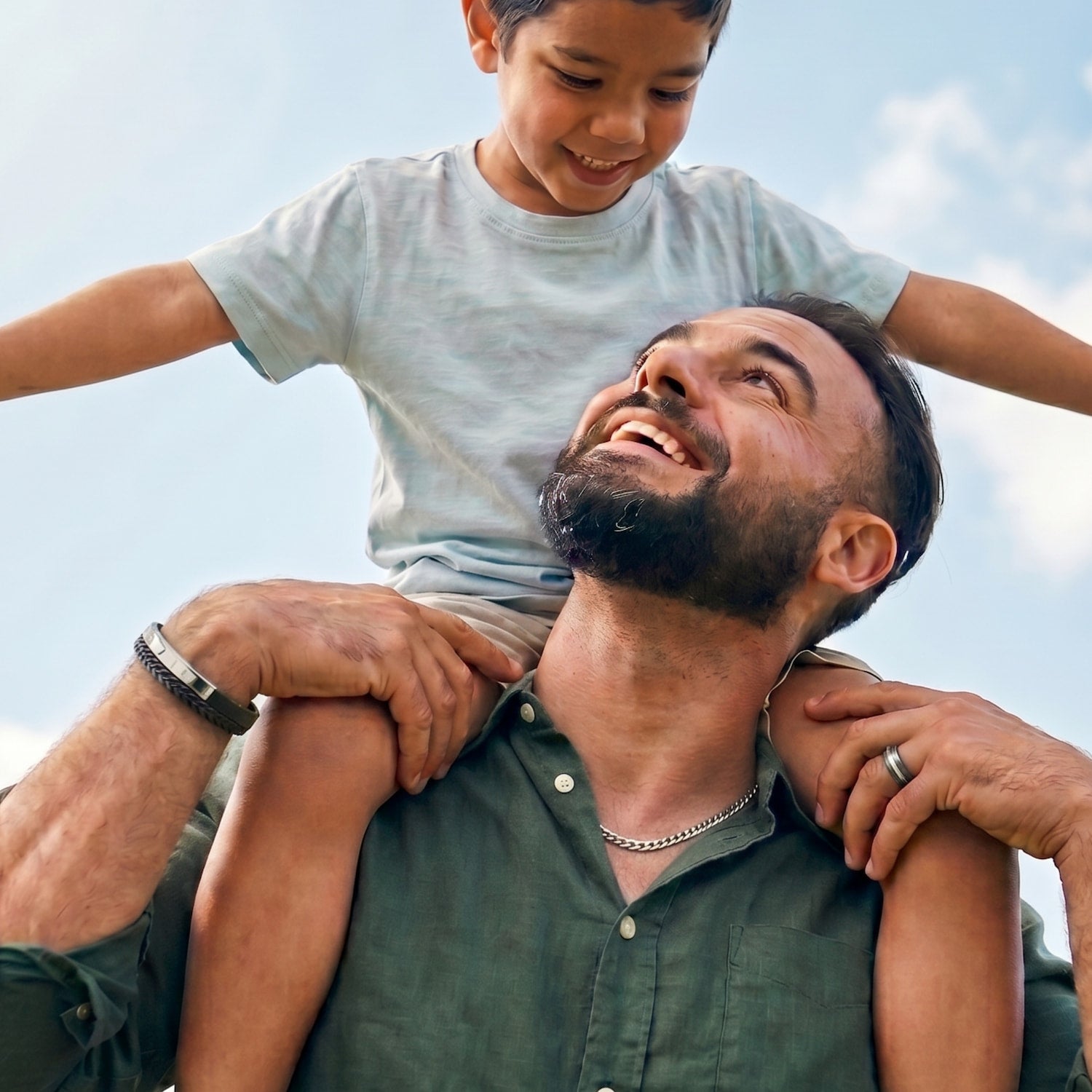 A smiling man with a beard wearing a silver chain necklace and a silver bracelet holds a young boy on his shoulders, both looking joyfully at each other against a bright sky.