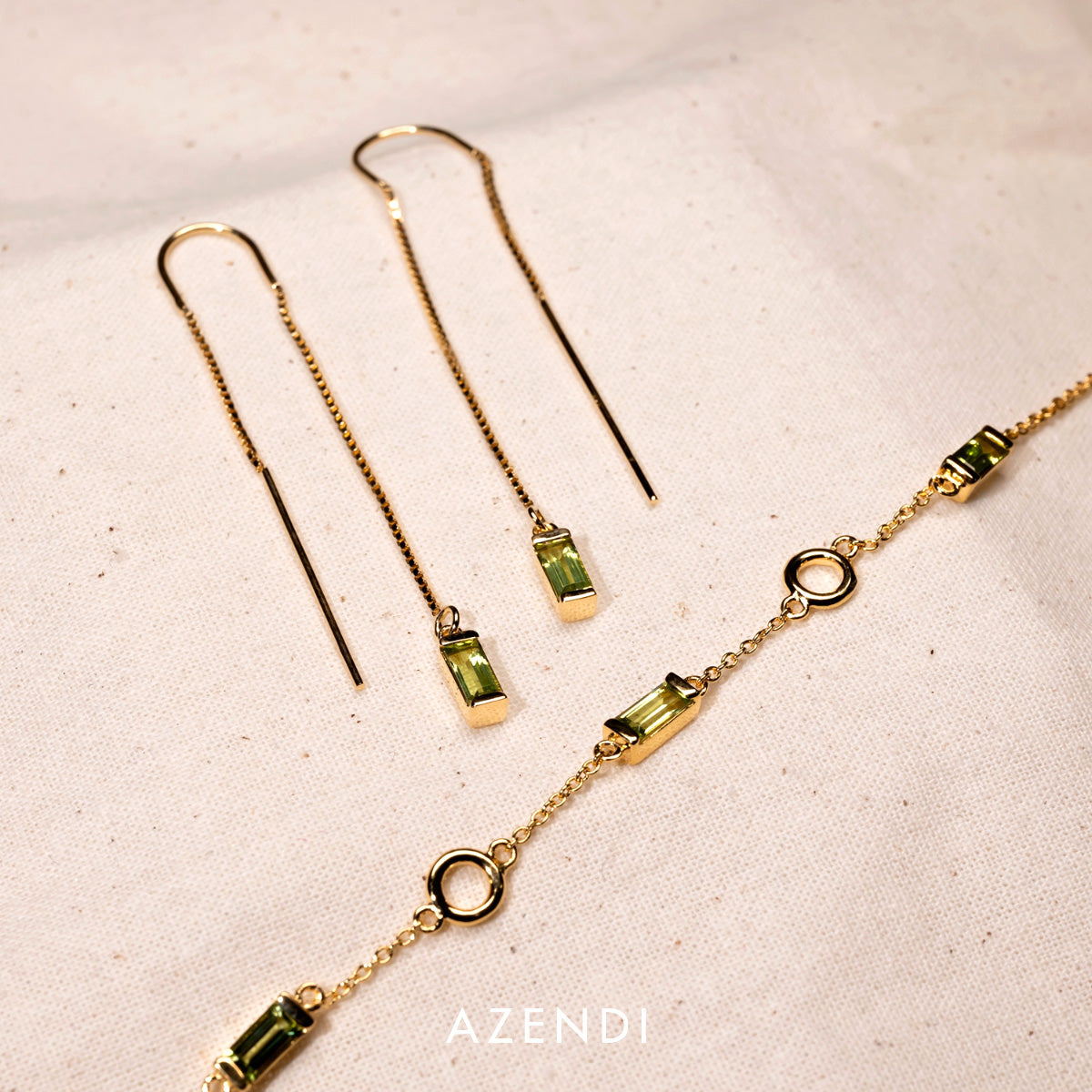 Delicate gold vermeil bracelet and matching threader earrings featuring rectangular peridot gemstones, displayed on a soft cream fabric background.