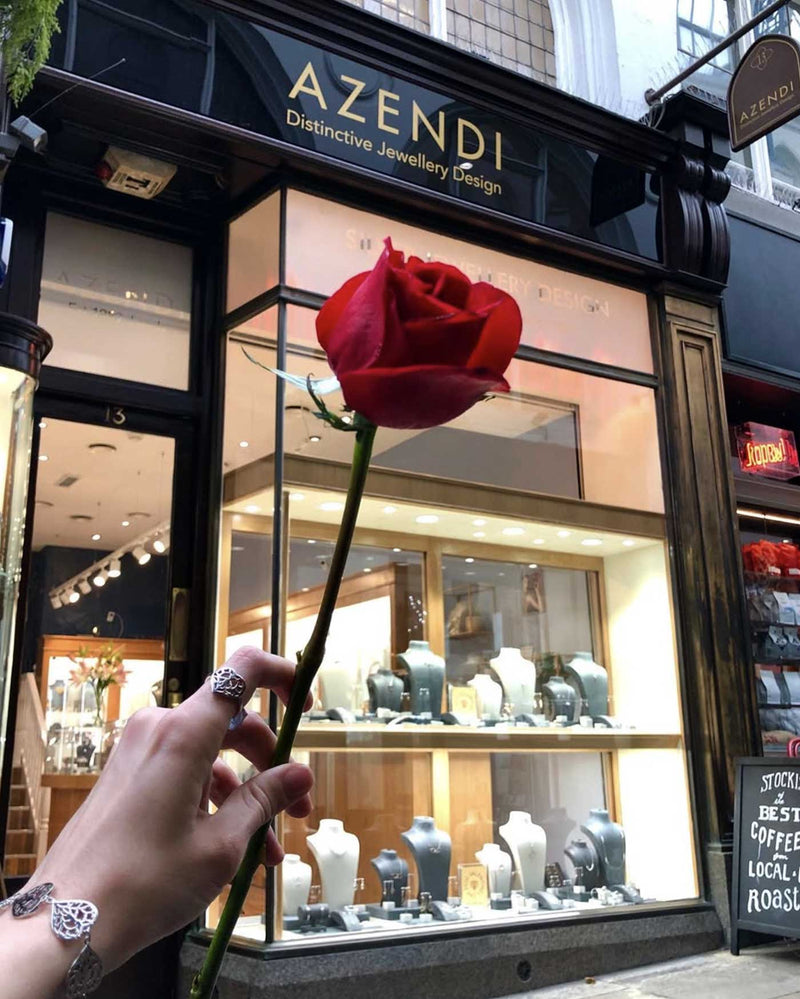 Hand holding a single red rose in front of a jewellery store window displaying a variety of sterling silver and gold vermeil pieces, with the store name prominently above.