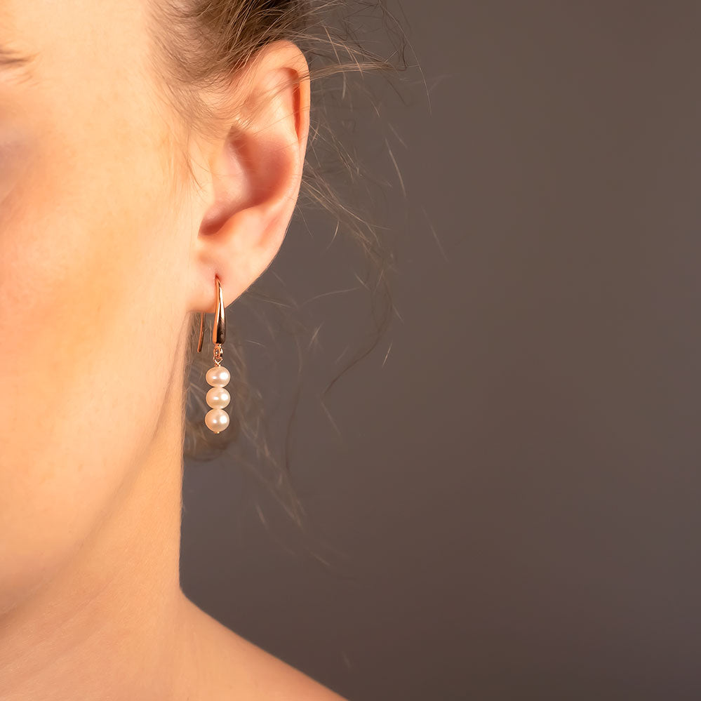 Close-up of an elegant earlobe wearing a delicate drop earring featuring three lustrous pearls suspended from a slender sterling silver hook against a soft neutral background.