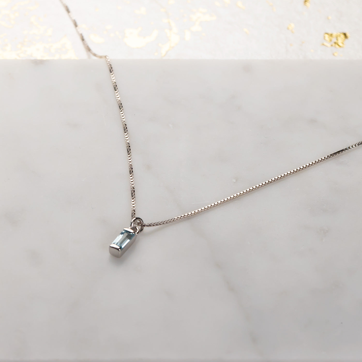 Delicate sterling silver necklace showcasing a slender baguette-cut blue topaz pendant with a polished bezel setting, resting on a smooth marble surface.