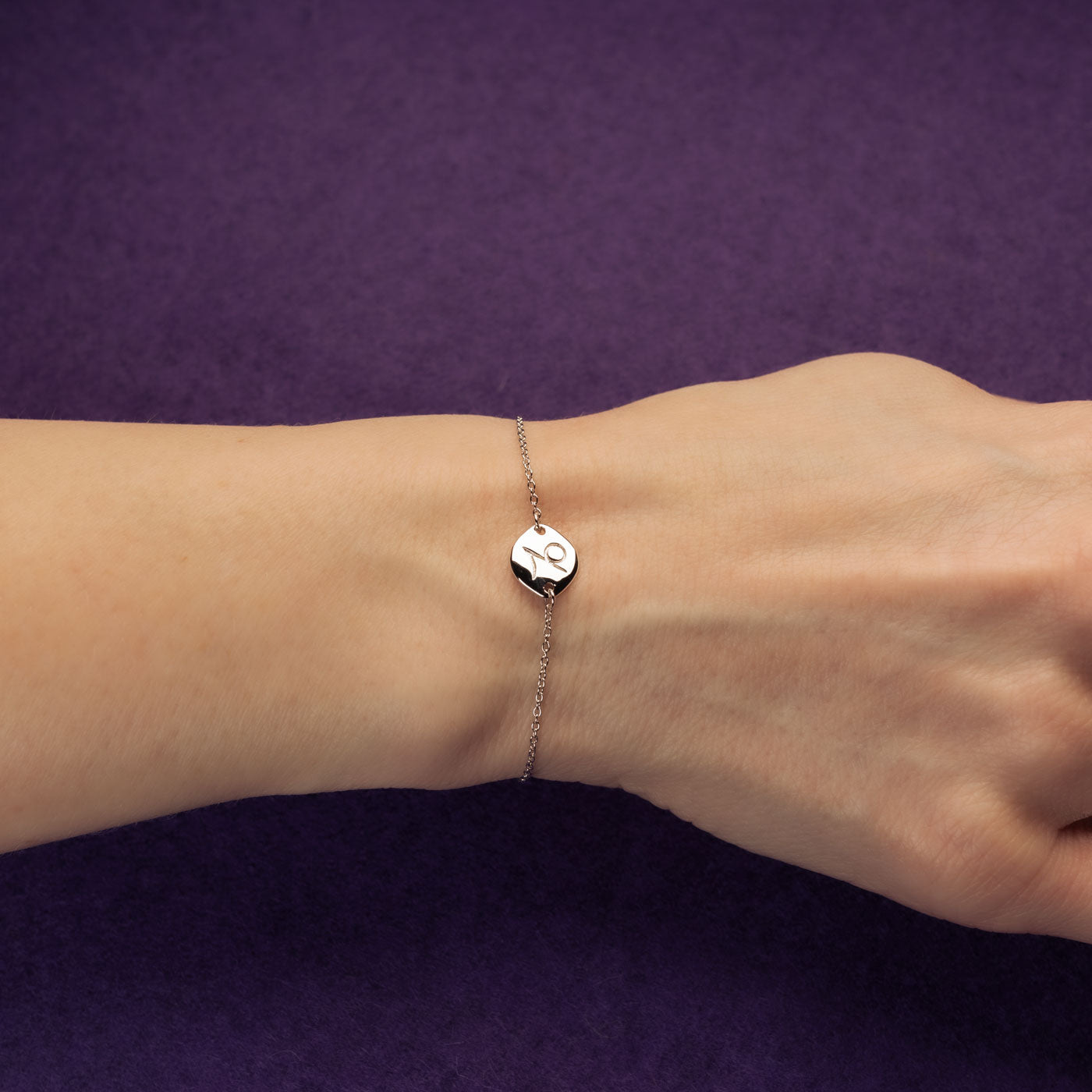 Delicate sterling silver bracelet featuring a Capricorn zodiac charm with an adjustable sliding bead, styled on a wrist against a deep purple background.