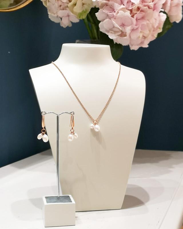 Delicate gold vermeil necklace and matching earrings featuring sculptural double freshwater pearl drops, displayed on minimalist white stands with soft pink hydrangeas in the background.